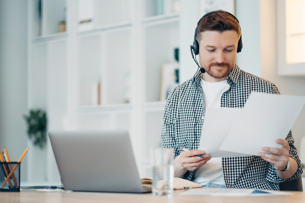 Das Bild zeigt einen Mann, der mit Headset am Laptop lernt und gerade Arbeitsunterlagen sichtet